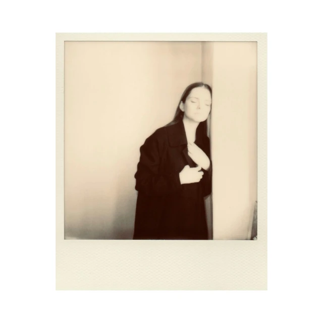 a black and white photo of a woman leaning against a wall, a polaroid photo, ( ( wearing a long coat ) ), on a pale background, cinestill!!, with instagram filters