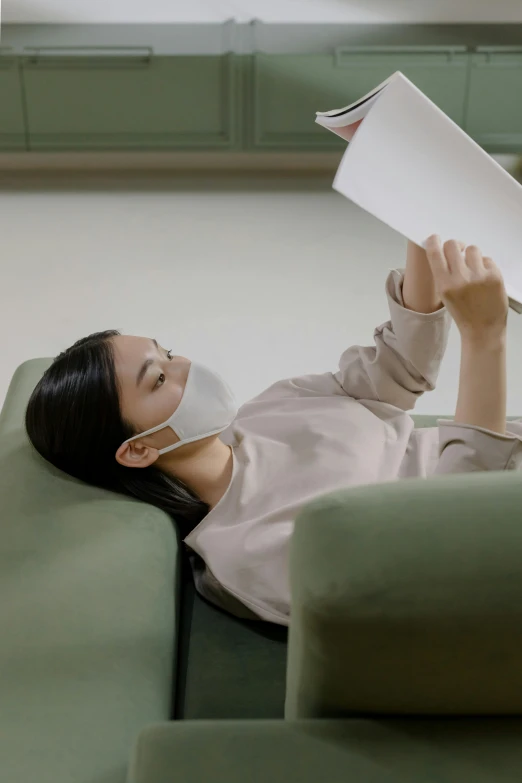 a woman laying on a couch reading a book, inspired by Fei Danxu, green facemask, sleek design, made from paper, ignant