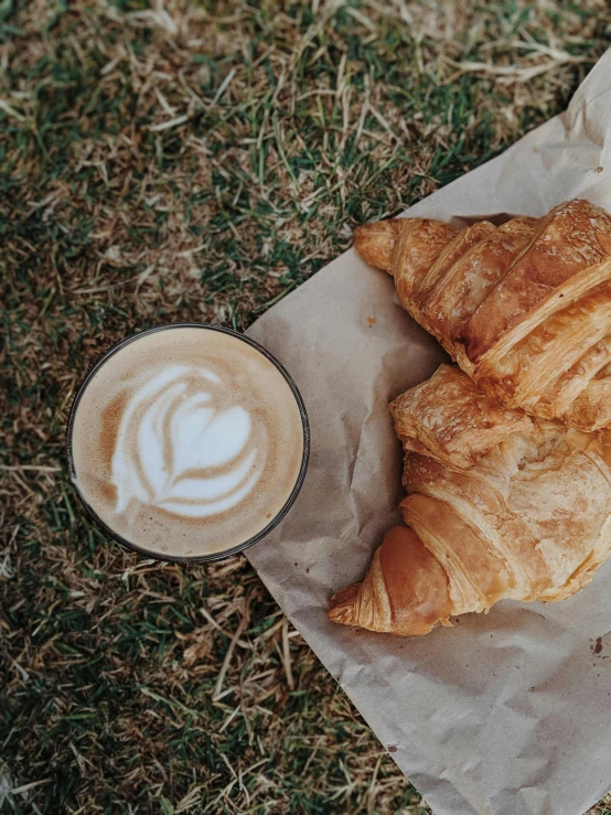 a couple of croissants sitting on top of a piece of paper next to a cup of coffee, by Austin English, picnic, 🚿🗝📝, iced latte, full frame image