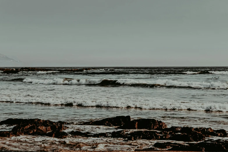 a man standing on top of a beach next to the ocean, by Carey Morris, pexels contest winner, minimalism, rough waves, muted browns, a photo of the ocean, background image