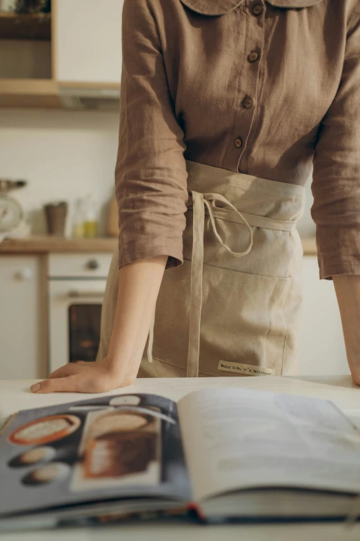 a woman reading a cookbook in a kitchen, inspired by Sarah Lucas, pexels contest winner, hyperrealism, white waist apron and undershirt, muted browns, wearing a linen shirt, gif