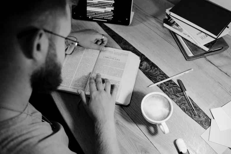 a man sitting at a table with a book and a cup of coffee, a black and white photo, by Mathias Kollros, pexels, bible, programming, maintenance photo, thumbnail