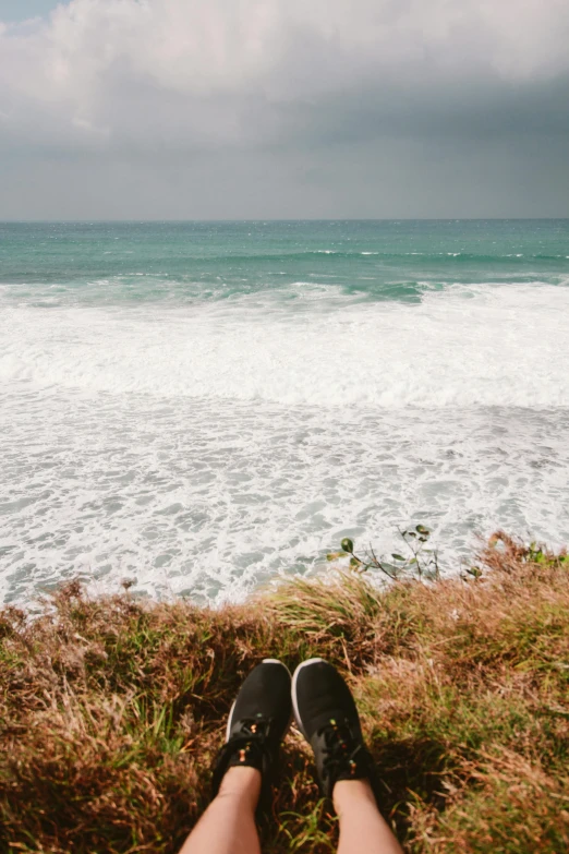 a person sitting on top of a grass covered hill next to the ocean, wet feet in water, bulli, cresting waves and seafoam, 2019 trending photo