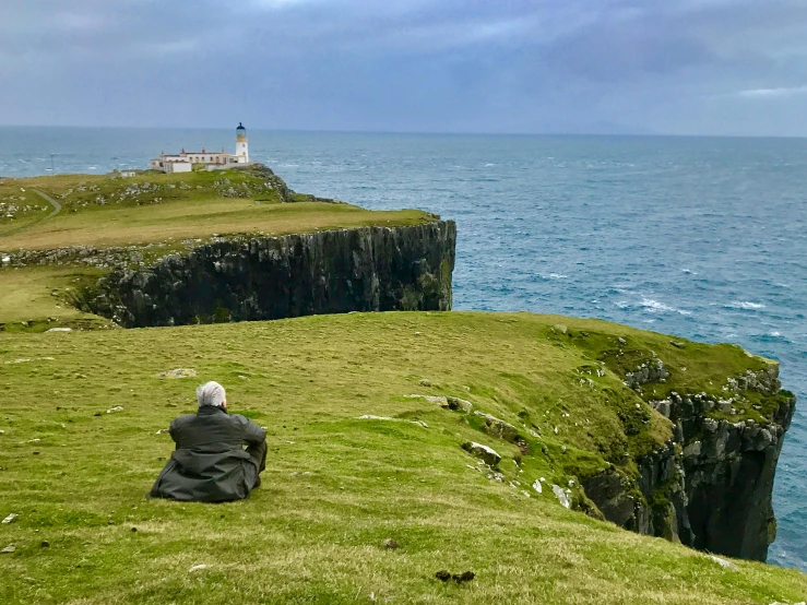 a man sitting on the edge of a cliff overlooking the ocean, by David Donaldson, pexels contest winner, old gigachad with grey beard, in a large grassy green field, bold lighthouse in horizon, skye meaker