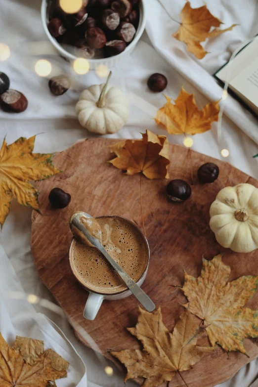 a cup of coffee sitting on top of a wooden cutting board, a still life, by Jessie Algie, pexels contest winner, glowing pumpkins under a tree, sitting on a mocha-colored table, dried leaves, gif
