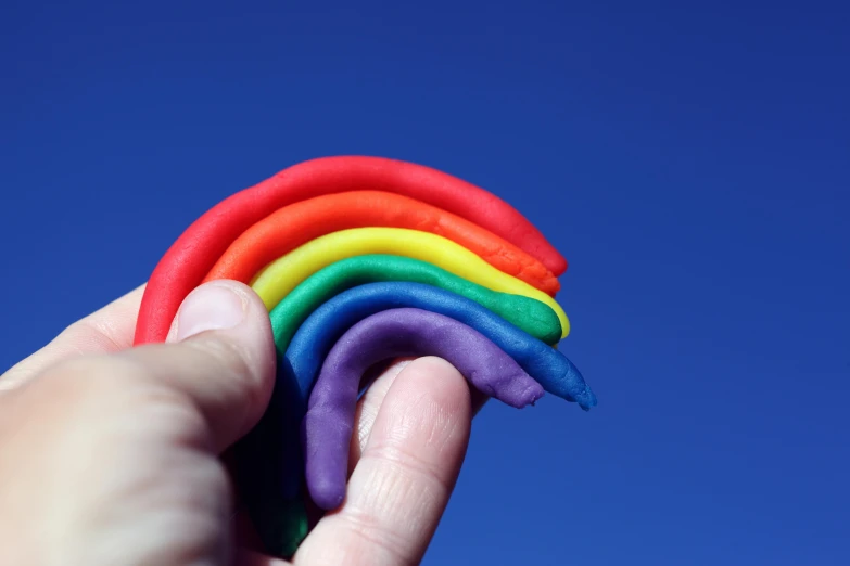 a hand holding a rainbow colored toy in front of a blue sky, unsplash, plasticien, plasticine, edible, on a hot australian day, homoerotic!!