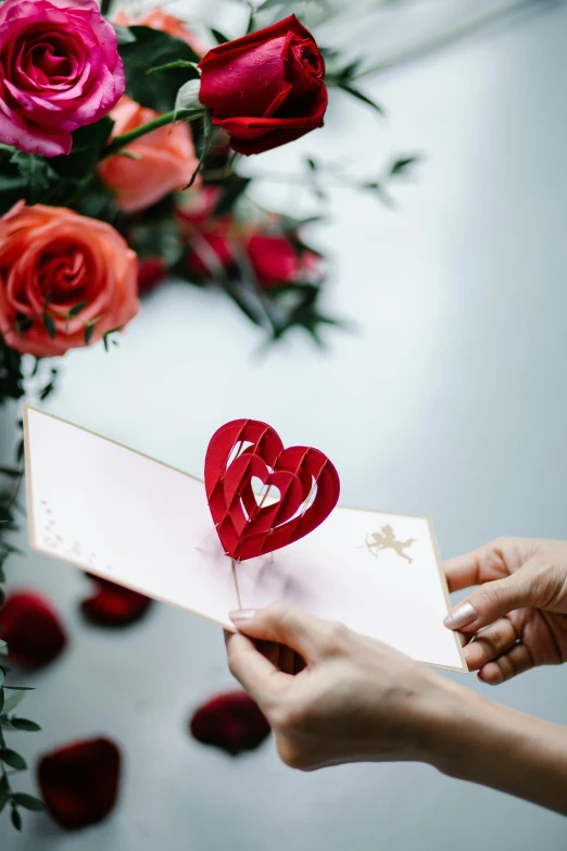 a person holding a card with a heart cut out of it, by Julia Pishtar, unsplash, exploding roses, kirigami, angled shot, greeting card