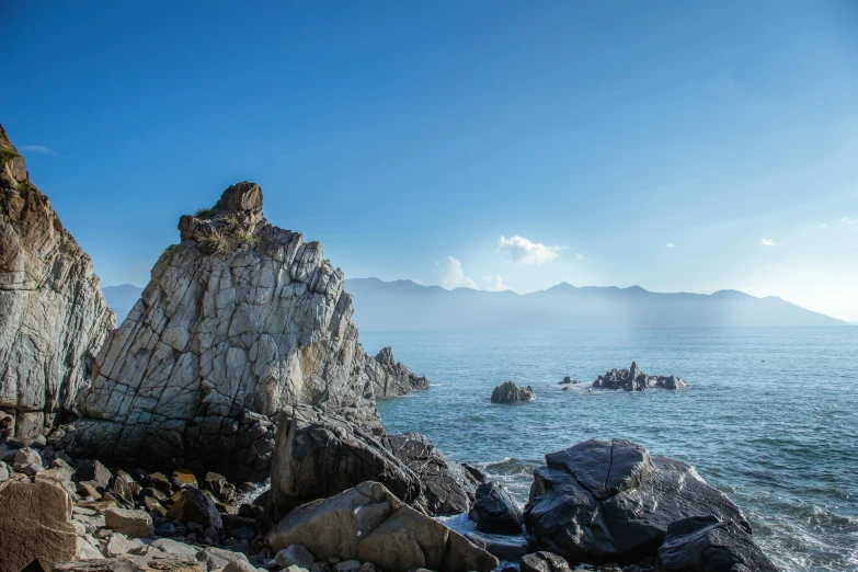 a group of rocks sitting on top of a rocky beach, a photo, mountains and ocean, profile image, andes, clear blue skies