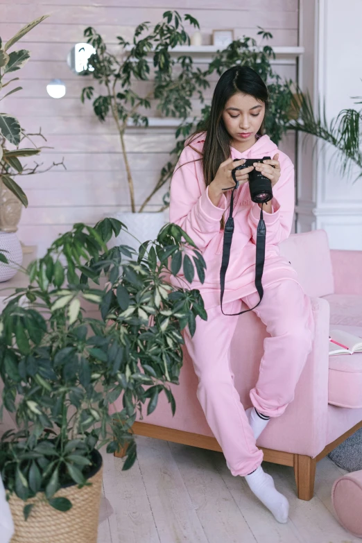 a woman sitting on a pink couch holding a camera, wearing a tracksuit, next to a plant, wearing a pastel pink hoodie, ((pink))