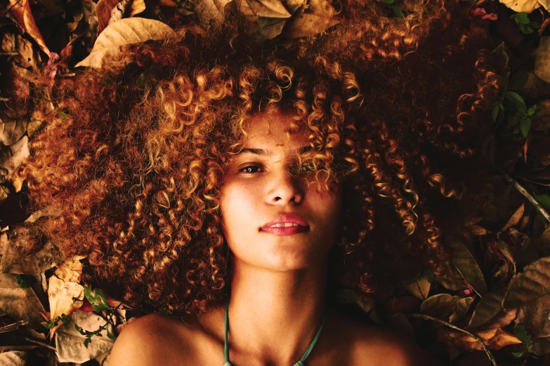 a woman laying on top of a pile of leaves, an album cover, trending on pexels, renaissance, curly copper colored hair, east african man with curly hair, high angle closeup portrait, natural skin tone 4k textures