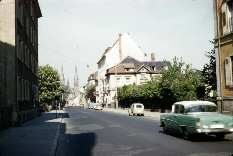 a green car driving down a street next to tall buildings, a colorized photo, by Werner Gutzeit, chartres cathedral, 1950s photograph, hasselblad photograph, album