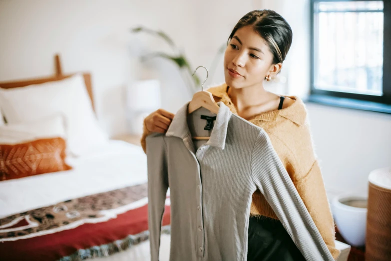 a woman standing in front of a bed holding a jacket, trending on pexels, inspect in inventory image, wearing a sweater, a young asian woman, wearing silver dress