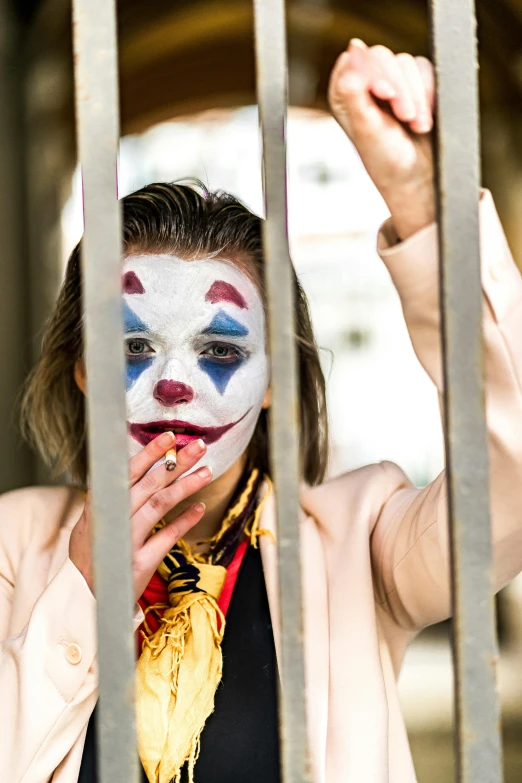 a woman with a clown face painted on her face behind bars, a portrait, by Julia Pishtar, trending on pexels, film still of batman, square, coloured, holding a cigarette