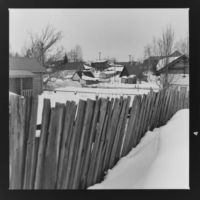 a black and white photo of a fence in the snow, by Maurycy Gottlieb, some houses in the background, ( ( photograph ) ), western town, ilya ozornin