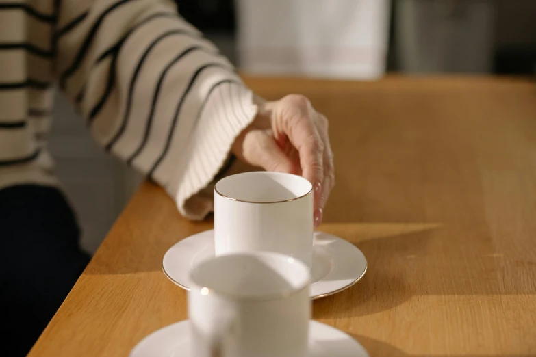 a person sitting at a table with a cup and saucer, up close, take my hand, carefully crafted, next to a cup