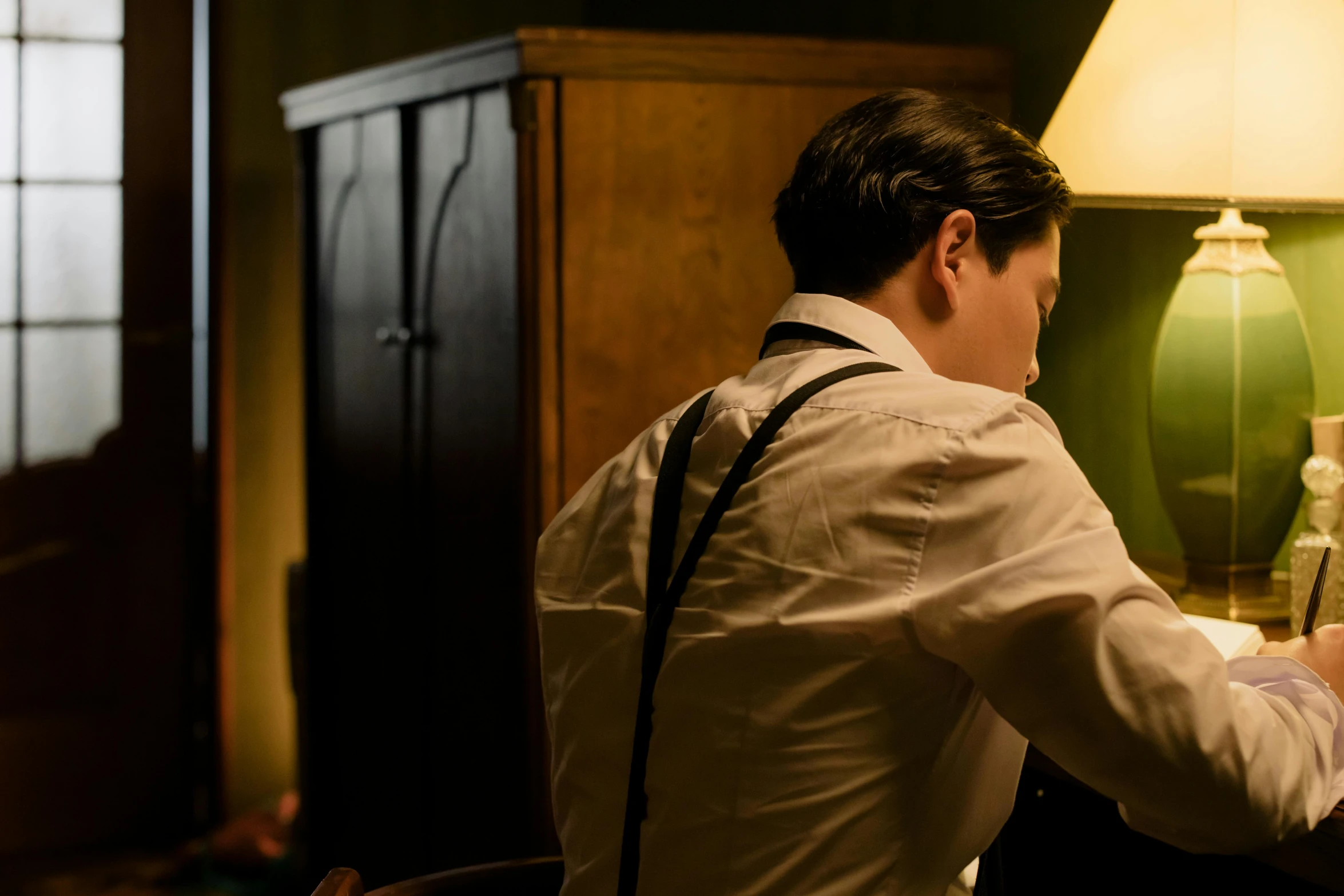 a man sitting at a desk in front of a lamp, inspired by Albert Edelfelt, unsplash, fantastic realism, still from loki ( 2 0 2 1 ), showing her shoulder from back, dressed like in the 1940s, jesper esjing