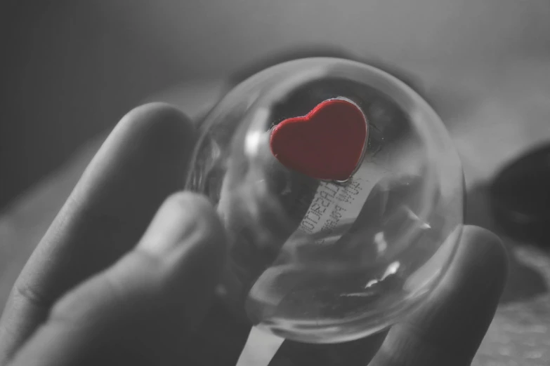 a person holding a glass ball with a red heart in it, a black and white photo, by Adam Marczyński, pexels, made of glass, just a cute little thing, coloured, love theme