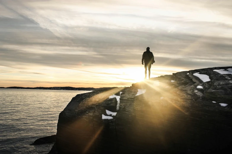 a man standing on top of a rock next to the ocean, by Jesper Knudsen, pexels contest winner, back lit, walking away, winter sun, archipelago