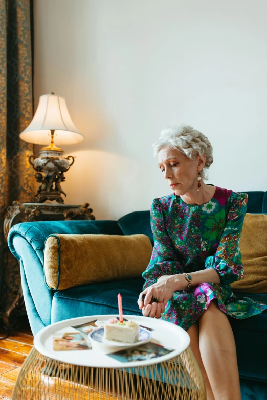 a woman sitting on a couch in a living room, a portrait, by Winona Nelson, holding a birthday cake, looking sad, elderly, marie - gabrielle capet style