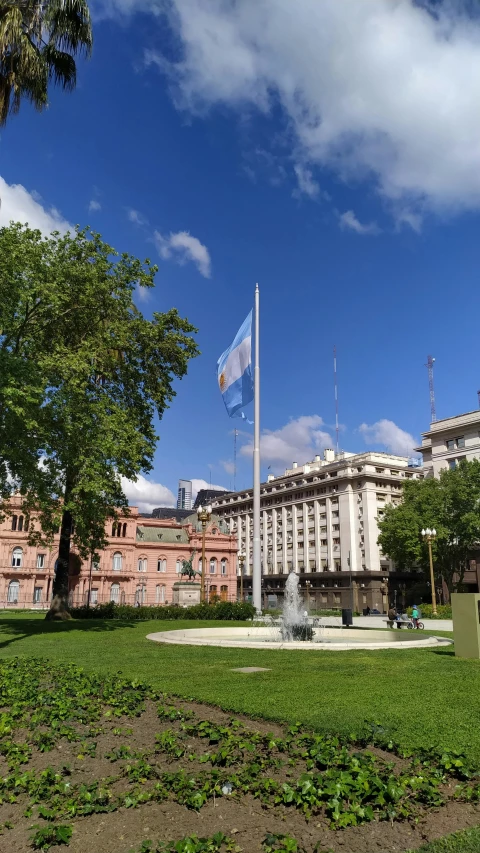 a large building sitting on top of a lush green field, inspired by Lajos Berán, flags of argentina behind, city square, light blue sky, 🚿🗝📝