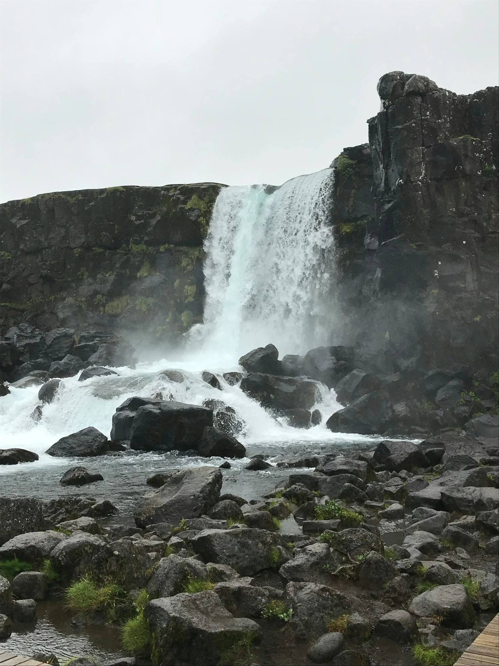 a man standing in front of a waterfall, an album cover, by Terese Nielsen, pexels contest winner, hurufiyya, with lots of dark grey rocks, viewed from a distance, reykjavik, very poor quality of photography