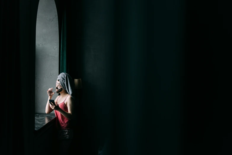a woman standing in front of a window looking at her cell phone, a portrait, inspired by Elsa Bleda, unsplash contest winner, behind red curtains, female death holding a cocktail, in an empty black room, hotel room