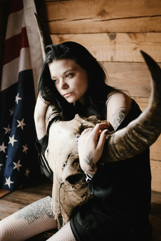 a woman sitting on top of a wooden floor next to an american flag, an album cover, unsplash, renaissance, black horns, bam margera, bone and ivory, headshot photo