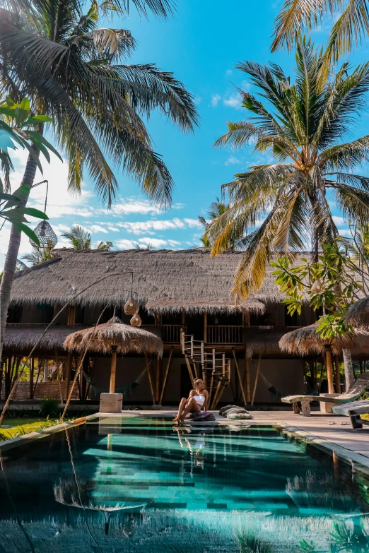 a person sitting on the edge of a swimming pool, bamboo huts, palm trees outside the windows, standing on surfboards, clear blue skies