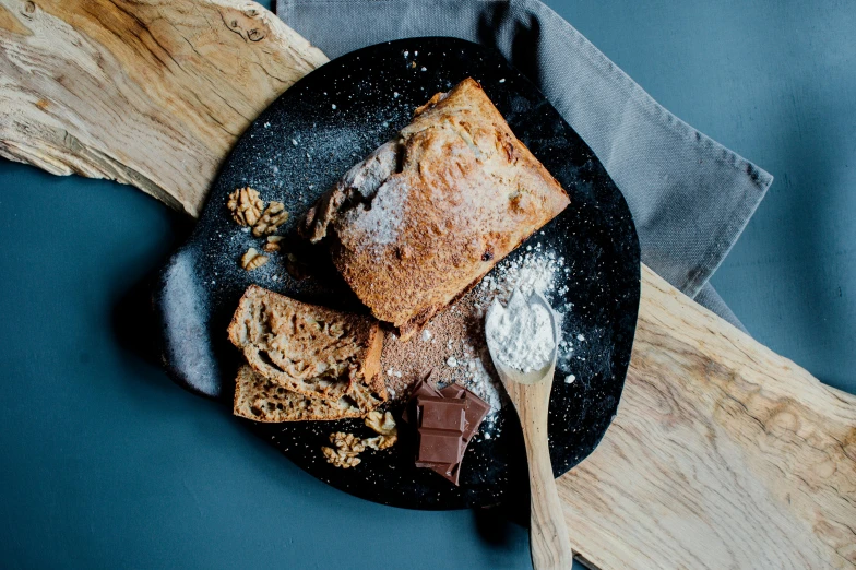 a piece of bread sitting on top of a black plate, unsplash, chocolate, with a wooden stuff, covered in white flour, various posed