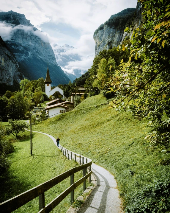 a person walking down a path next to a lush green hillside, inspired by Karl Stauffer-Bern, pexels contest winner, romanesque, snow capped mountains, churches, conde nast traveler photo, 7 0 s photo