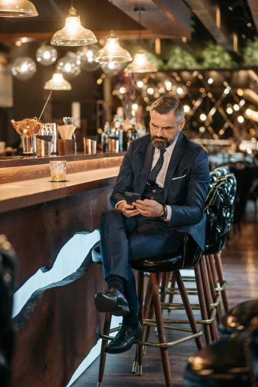 a man sitting at a bar looking at his phone, happening, wearing a stylish men's suit, connectivity, full body image, insightful