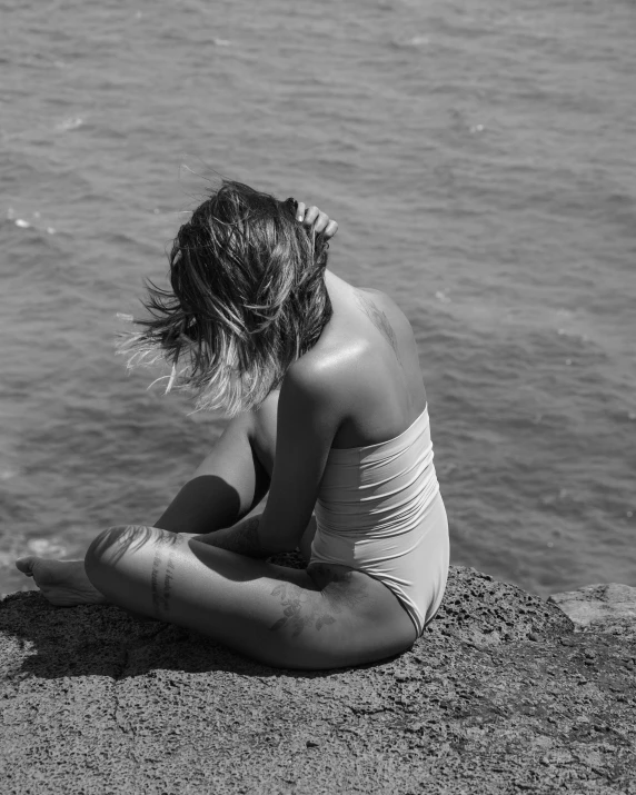 a woman sitting on top of a beach next to a body of water, a black and white photo, inspired by Max Dupain, unsplash, wearing white leotard, girl with messy bun hairstyle, in the sun, sad feeling