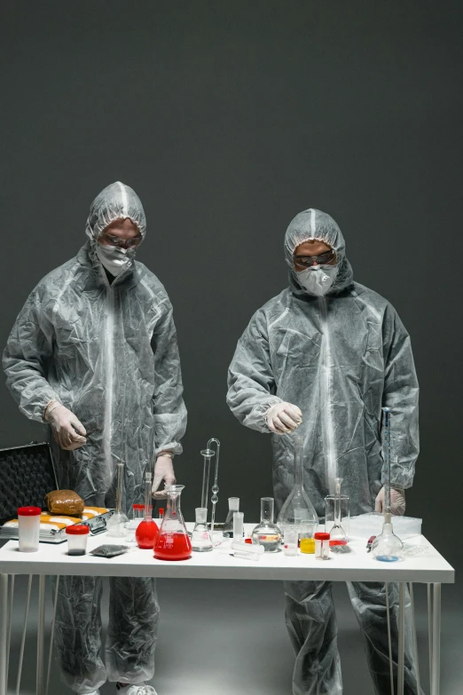 a couple of people that are standing in front of a table, by Adam Marczyński, staff wearing hazmat suits, on grey background, taking mind altering drugs, plating