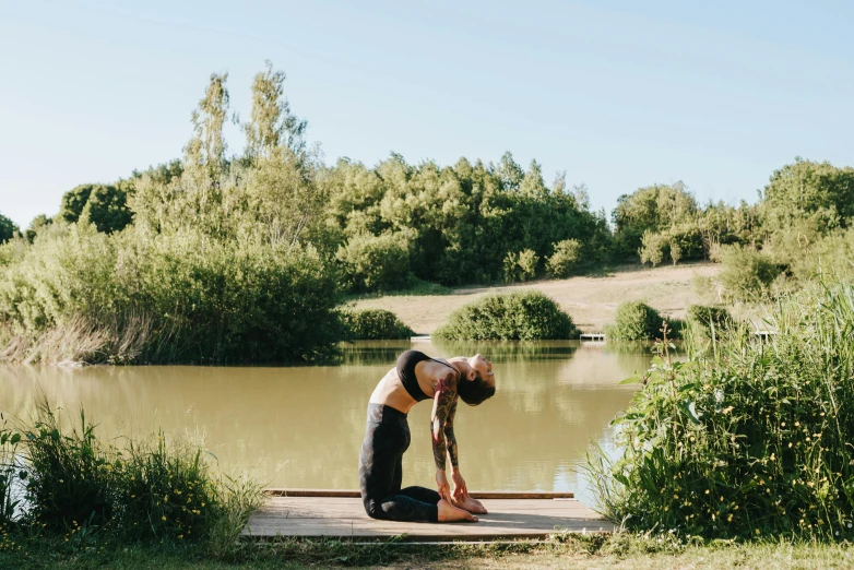 a woman doing a yoga pose in front of a body of water, by Arabella Rankin, unsplash, process art, in a park and next to a lake, back arched, manuka, near pond