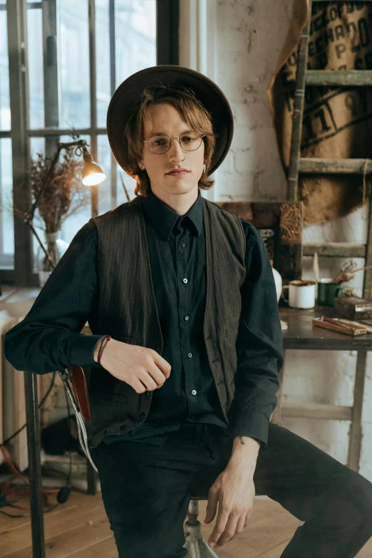 a man sitting on a chair wearing a hat, an album cover, inspired by August Sander, trending on pexels, renaissance, black vest, rounded eyeglasses, portrait androgynous girl, indoor scene