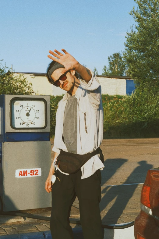 a man standing in front of a gas station, an album cover, unsplash, bauhaus, waving, low quality photo, very sunny, liam