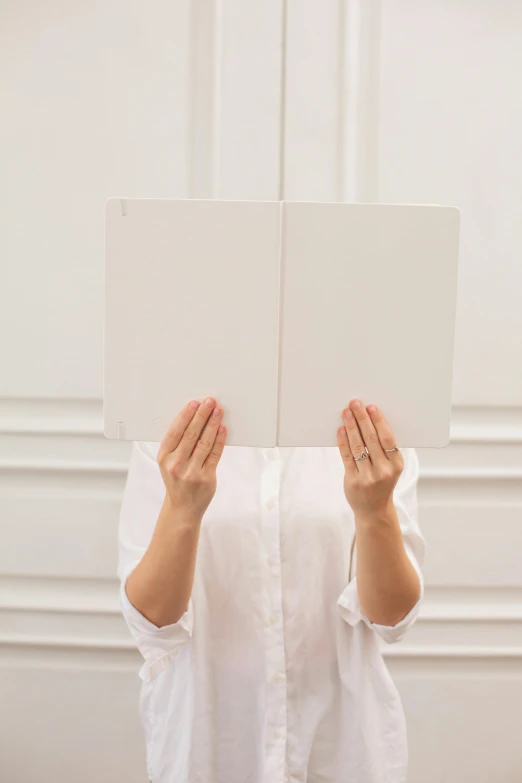 a woman covering her face with a sheet of paper, a minimalist painting, by Carey Morris, unsplash, he is holding a large book, wearing a white button up shirt, left right symmetry, made of all white ceramic tiles