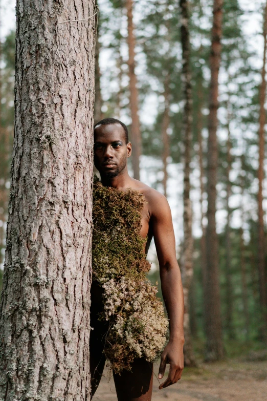 a man standing next to a tree in a forest, an album cover, inspired by Oluf Høst, wearing loincloth, photographed for reuters, brown skinned, finnish naturalism