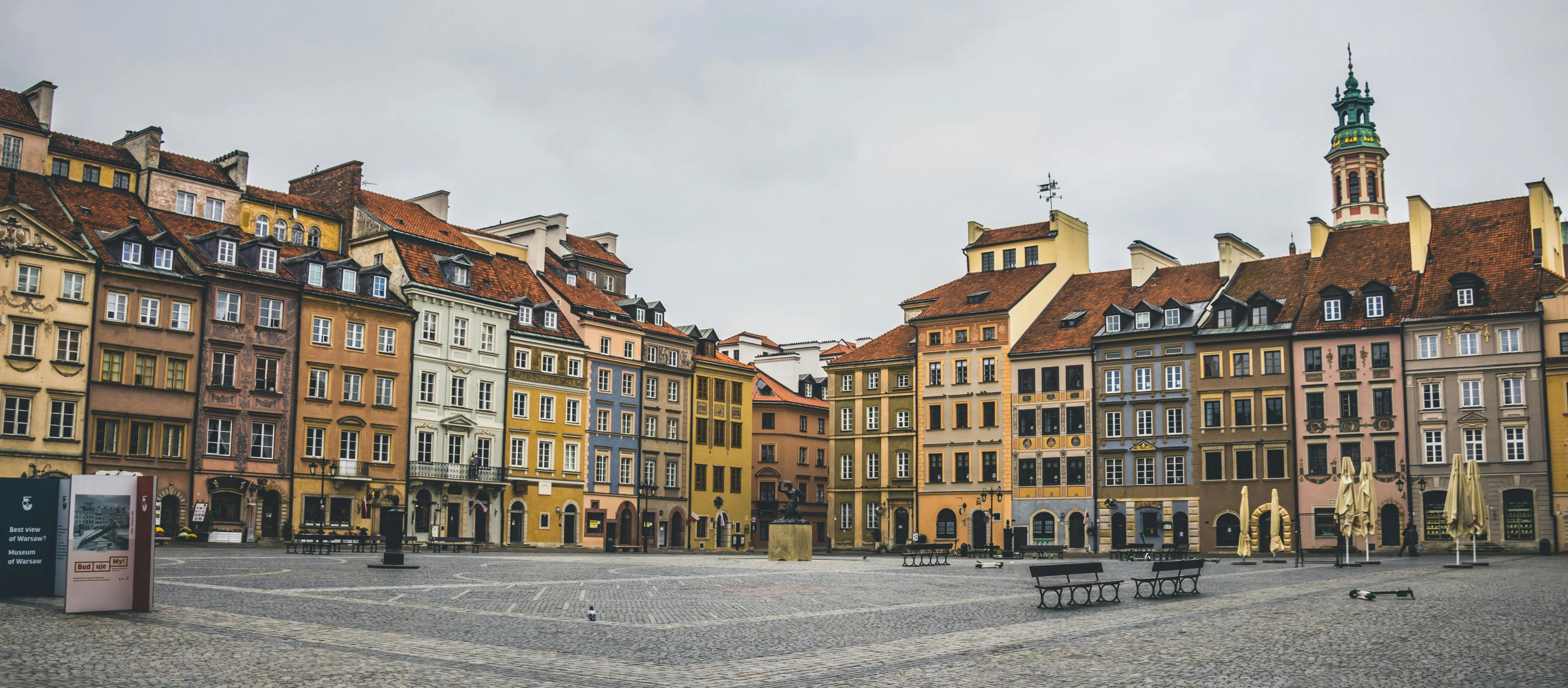 a couple of benches sitting on top of a cobblestone street, by Adam Marczyński, pexels contest winner, colorful building, daniel libeskind, in a square, full building