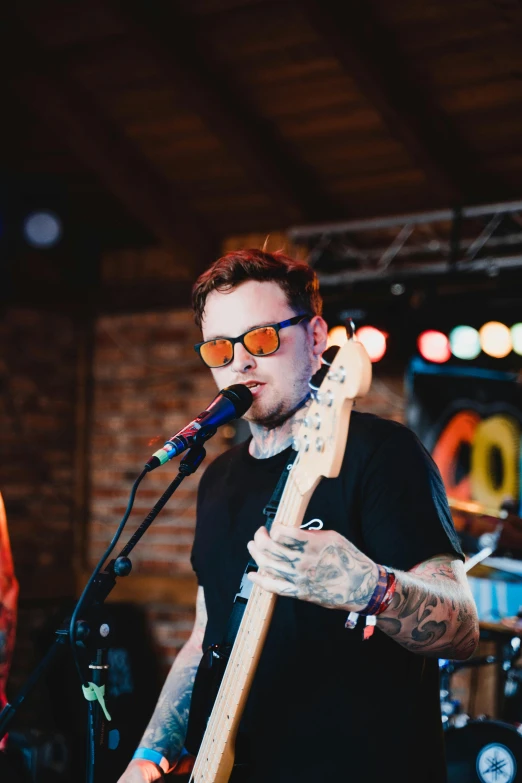 a man holding a guitar while standing in front of a microphone, inspired by Seb McKinnon, wearing orange sunglasses, profile image, with tattoos, bassist