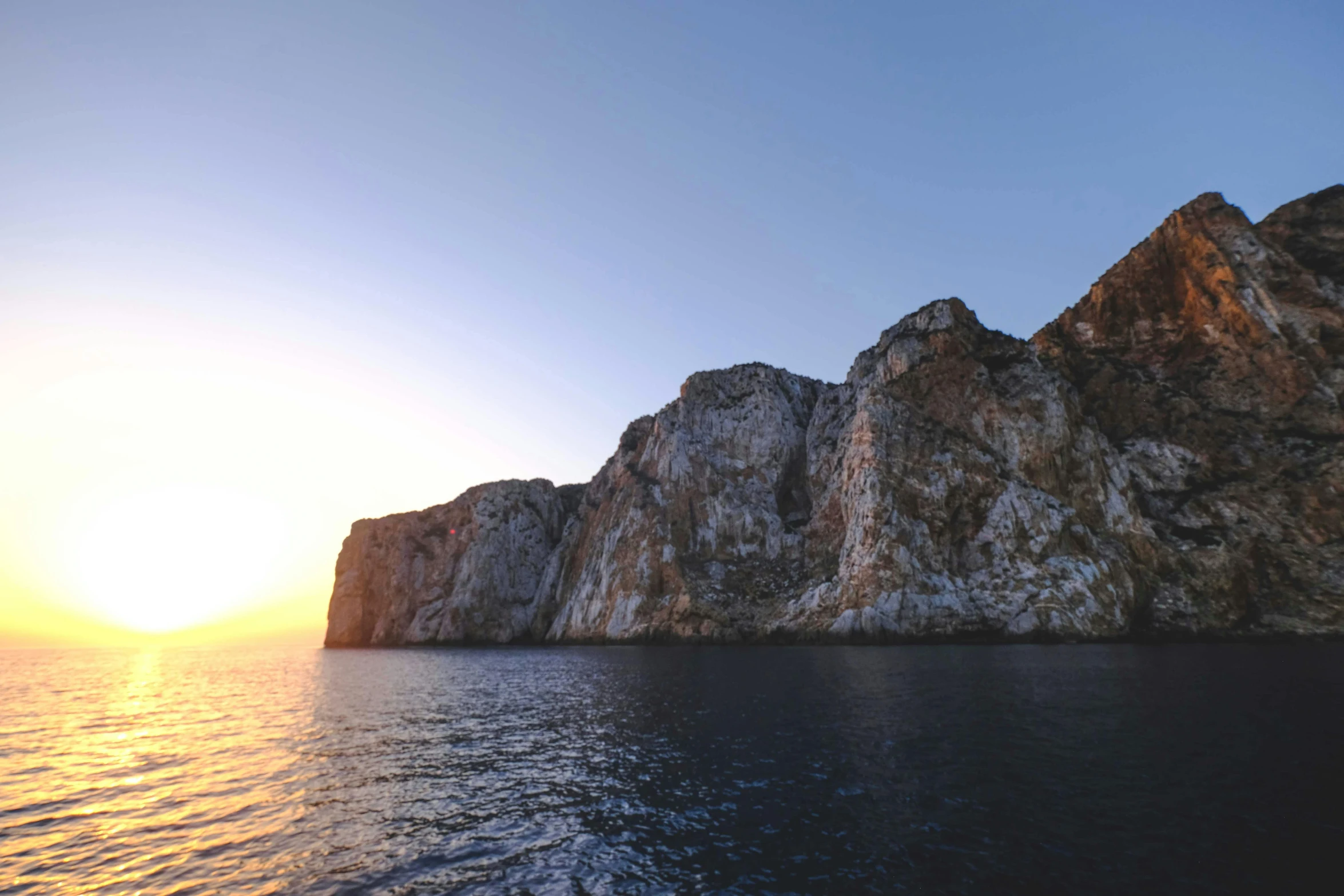 a large rock in the middle of a body of water, a digital rendering, pexels contest winner, romanticism, mediterranean vista, 8k hdr sunset lit, steep cliffs, photo taken from a boat