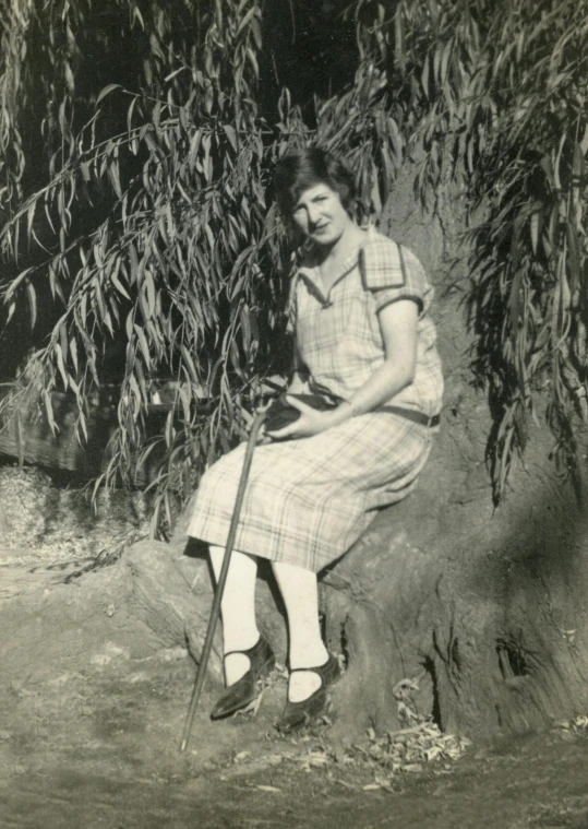 a black and white photo of a woman sitting under a tree, holding a cane, early 20s, digital image, museum photograph