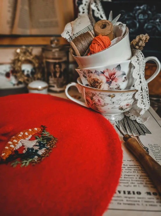 a red hat sitting on top of a table next to a cup, a cross stitch, inspired by Carl Larsson, pexels contest winner, vintage aesthetic, product introduction photo, 🤠 using a 🖥, an oversized beret