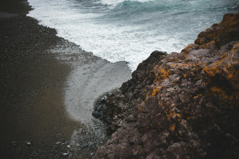 a man standing on top of a rocky beach next to the ocean, an album cover, unsplash contest winner, dark grey and orange colours, wave of water particles, covered in coral and barnacles, black sand