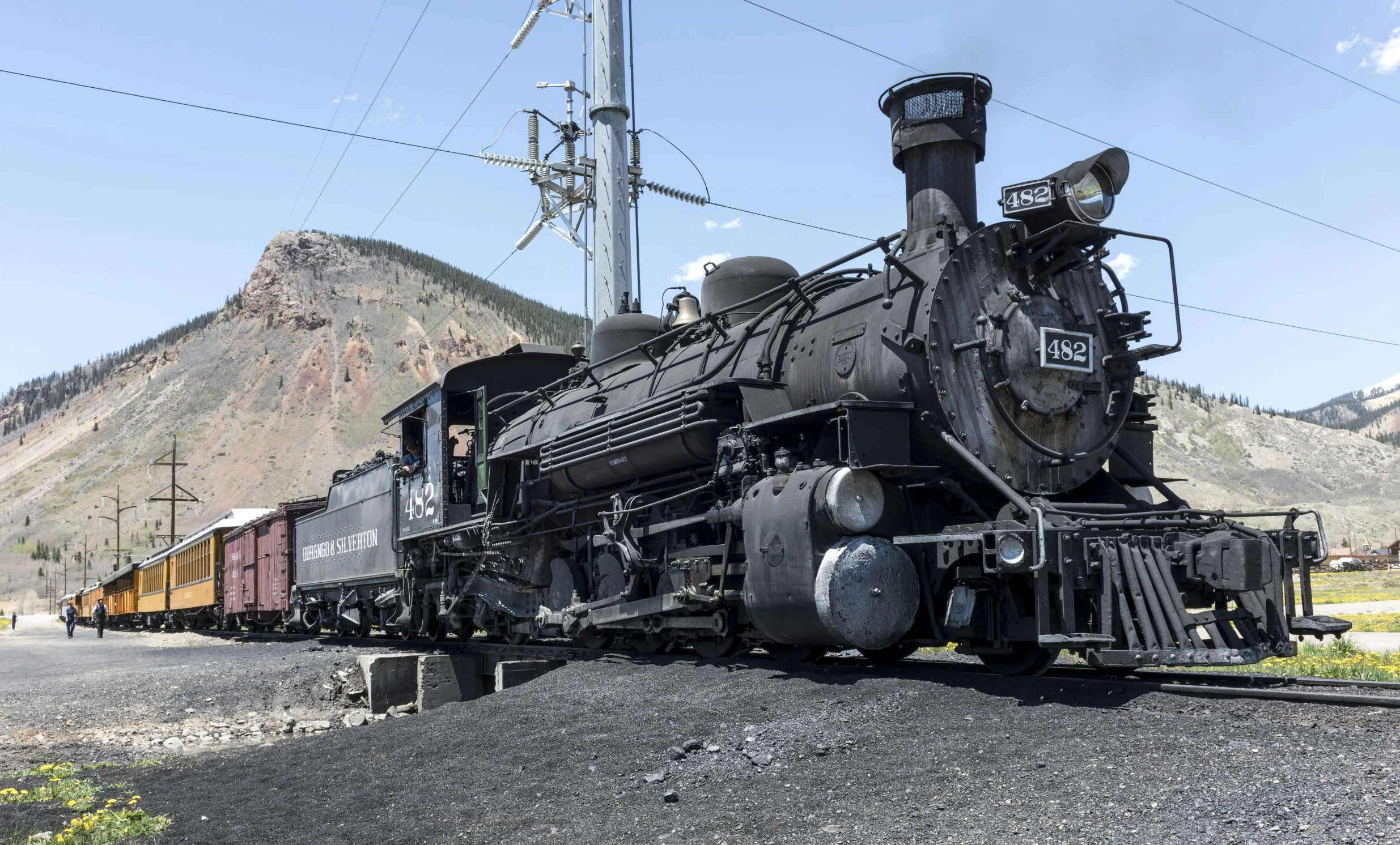 a train traveling down train tracks next to a mountain, featuring engine, one black, preserved historical, promo image