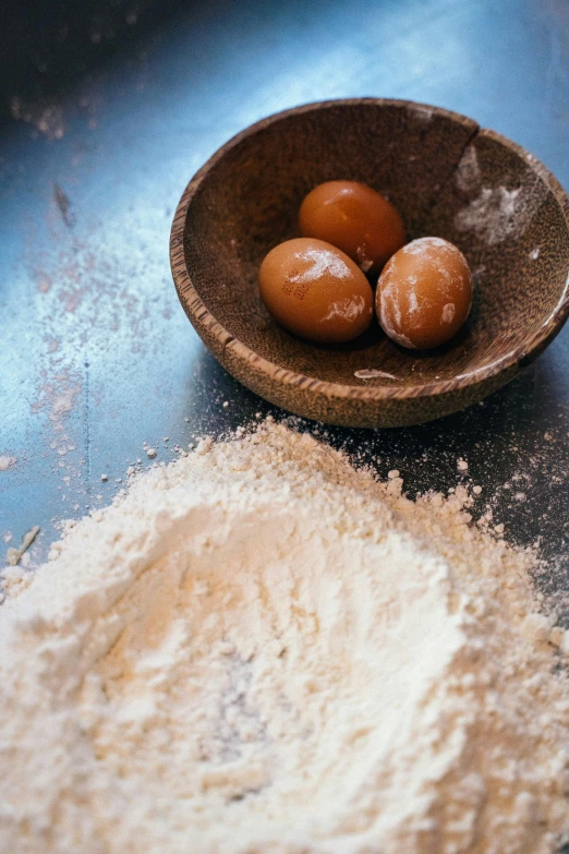 a wooden bowl filled with doughnuts next to a pile of flour, a still life, unsplash, renaissance, an egg, square, medium format. soft light, moroccan