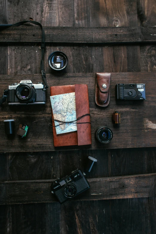 a wooden table topped with a camera and a notebook, items, map, model posing, looking down on the camera