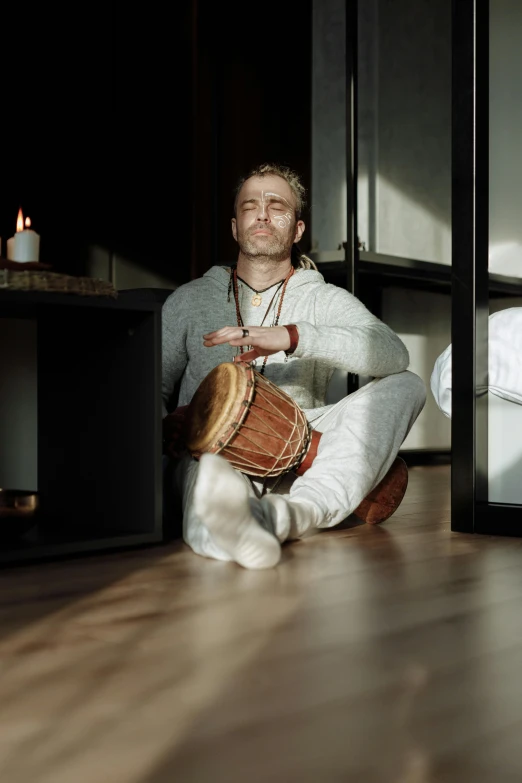 a man sitting on the floor with a drum, inspired by Samu Börtsök, unsplash, renaissance, meditating pose, upright, at home, hugging his knees