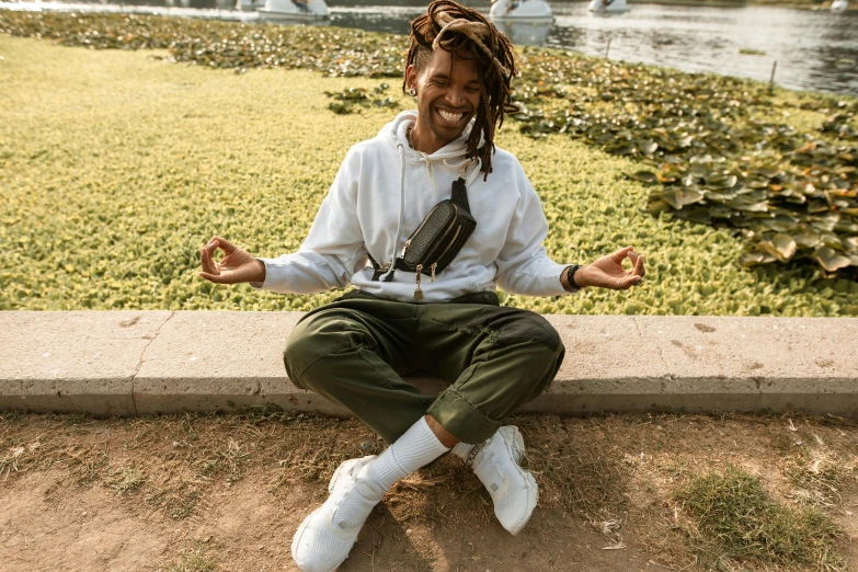 a man with dreadlocks sitting in front of a body of water, willow smith young, taking control while smiling, sukhasana, in a city park