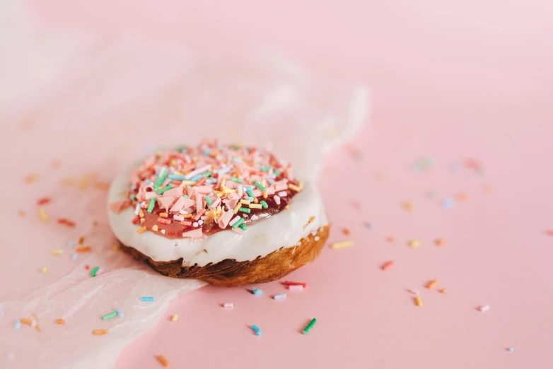 a doughnut with frosting and sprinkles on a pink surface, by Emma Andijewska, trending on unsplash, white and pink cloth, background image, desserts, 🥥 🍕 hybrid